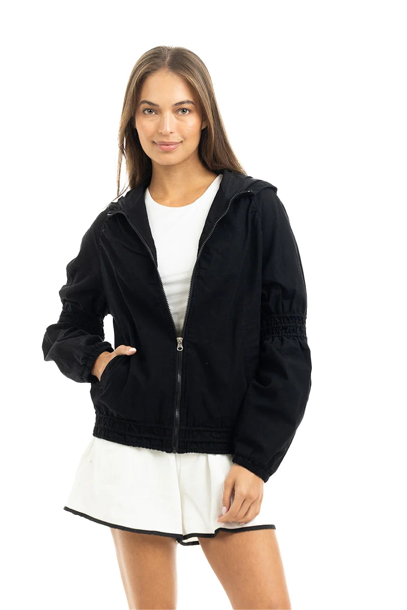 A woman with long brown hair smiles slightly as she models the Katydid Black Lightweight Hooded Cute Jacket over a white flared dress, standing against a plain white background.