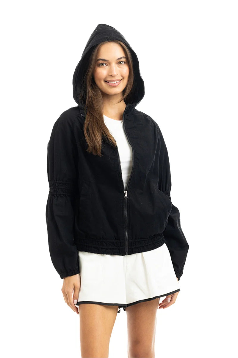 A woman models the Katydid Black Lightweight Hooded Cute Jacket over a white dress, smiling at the camera against a plain white background.
