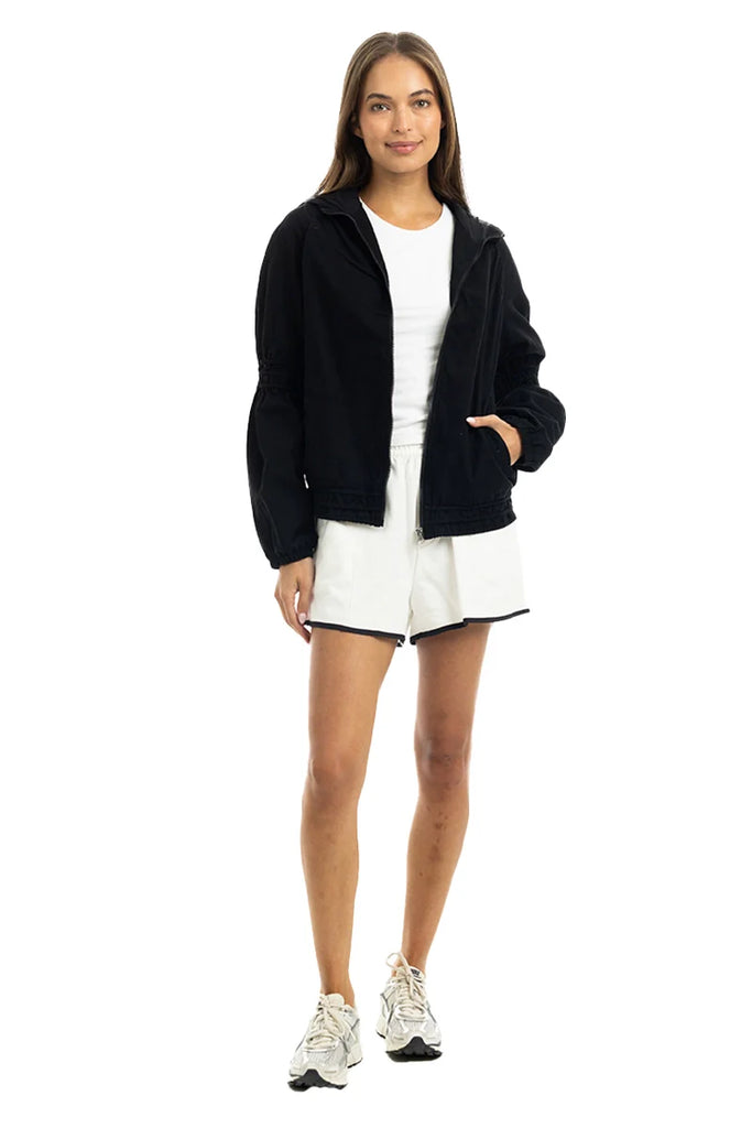 A woman poses confidently in a Katydid Black Lightweight Hooded Cute Jacket, paired with a white t-shirt, white shorts with black trim, and white sneakers. She stands against a white background with one hand in her pocket and long hair down.