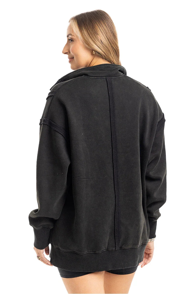 A person with light brown hair faces away, wearing the Katydid Black Vintage Wash Pullover Sweatshirt, which has visible seams down the back and shoulders, paired with black shorts. The background is plain white.