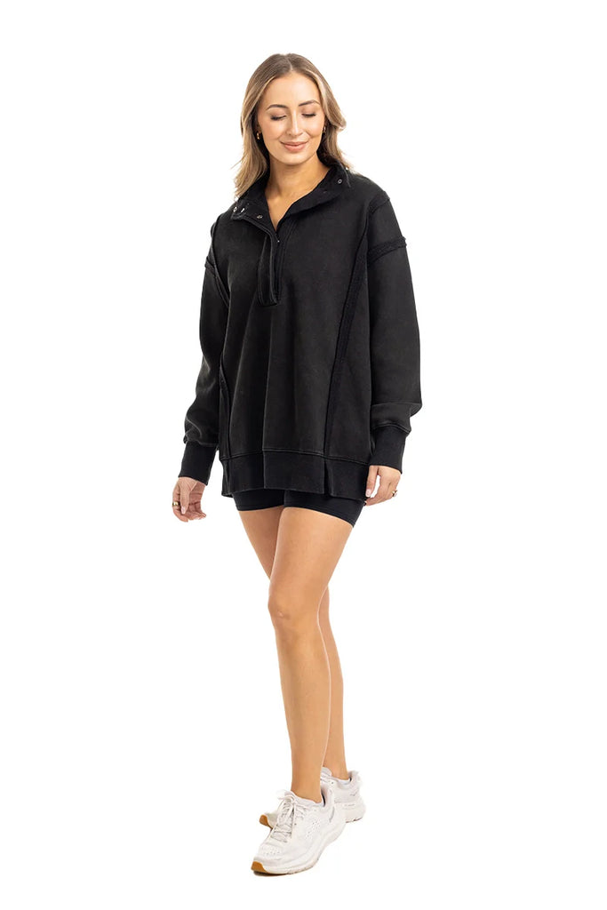 A woman smiles in a Katydid Black Vintage Wash Pullover Sweatshirt, styled oversized with black shorts and white sneakers, set against a plain white background.