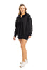 A woman smiles in a Katydid Black Vintage Wash Pullover Sweatshirt, styled oversized with black shorts and white sneakers, set against a plain white background.