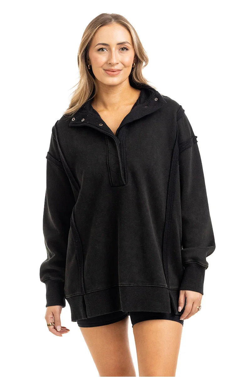 A woman with long, light brown hair smiles in a Katydid Black Vintage Wash Pullover Sweatshirt, featuring a short zipper. She pairs it with matching black shorts and stands against a plain white background.