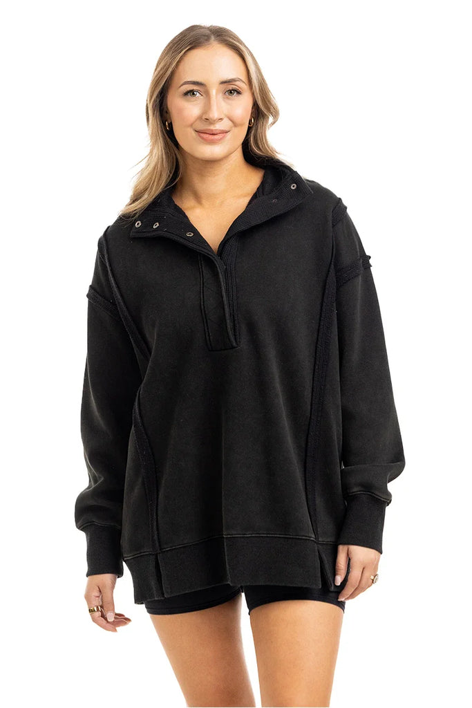 A woman with long, light brown hair smiles in a Katydid Black Vintage Wash Pullover Sweatshirt, featuring a short zipper. She pairs it with matching black shorts and stands against a plain white background.