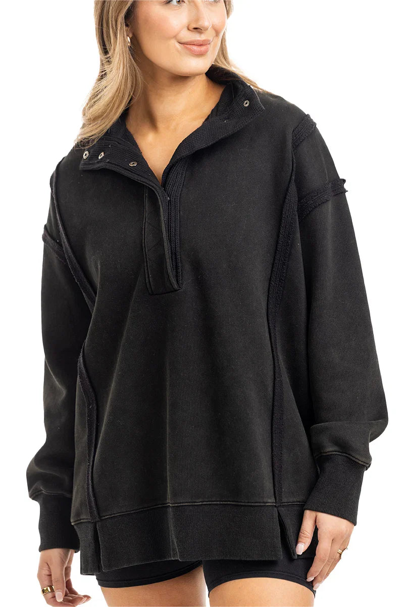 A woman wears the Katydid Black Vintage Wash Pullover Sweatshirt, featuring a loose fit, partial button-up collar, and long sleeves, standing with one hand relaxed by her side.