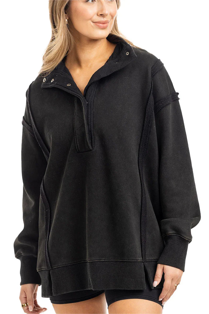 A woman wears the Katydid Black Vintage Wash Pullover Sweatshirt, featuring a loose fit, partial button-up collar, and long sleeves, standing with one hand relaxed by her side.