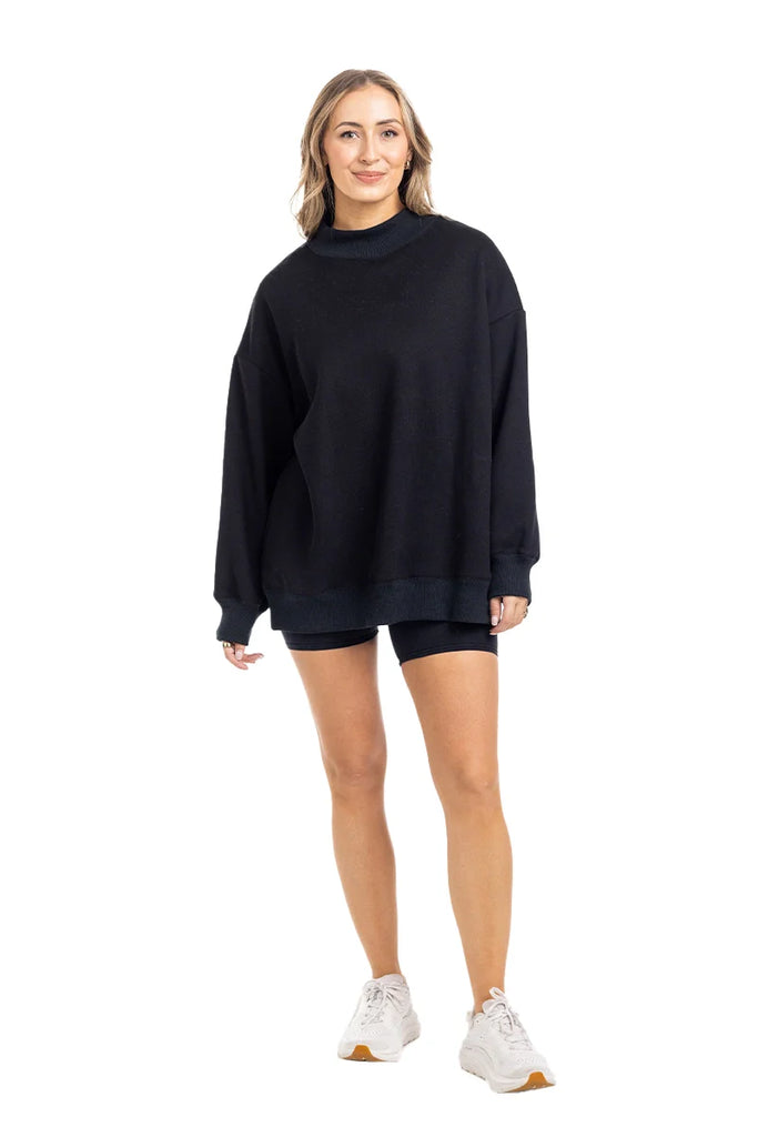 A woman with shoulder-length blonde hair smiles while wearing the Katydid Black Mockneck Sweatshirt, black shorts, and white sneakers, posing against a plain white background.