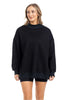 A woman with light brown hair is wearing the Katydid Black Mockneck Sweatshirt and black shorts, standing and smiling against a white background.