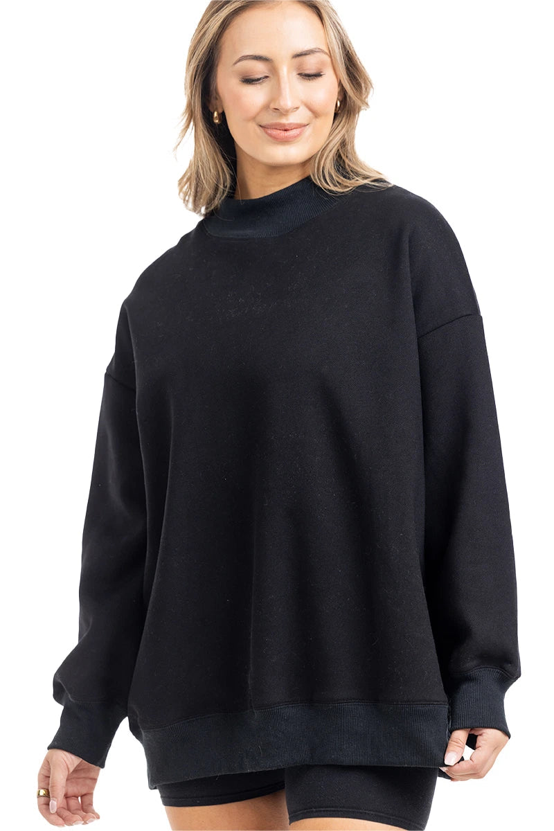 A woman with shoulder-length blonde hair smiles with her eyes closed, wearing the Katydid Black Mockneck Sweatshirt and black shorts against a white background.