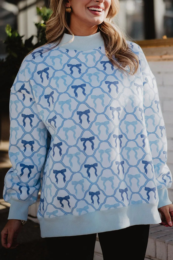 A woman smiles outdoors wearing the Katydid Light Blue Mini Bows Mockneck Sweatshirt—a cozy, loose-fitting, long-sleeve top in light blue featuring a repeating mini bows pattern.