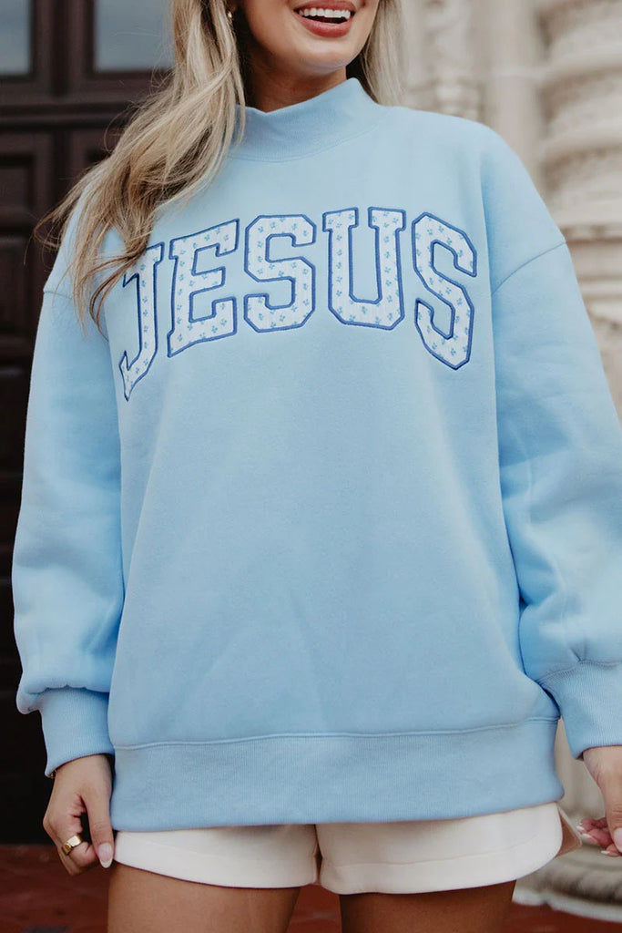 A woman outdoors smiles in a faith-inspired style, wearing white shorts and the Katydid JESUS Mockneck—a light blue sweatshirt with large, bold JESUS lettering on the front.