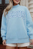 A woman outdoors smiles in a faith-inspired style, wearing white shorts and the Katydid JESUS Mockneck—a light blue sweatshirt with large, bold JESUS lettering on the front.