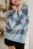 A smiling person indoors showcases the Katydid Light Blue and Navy Striped Mockneck Sweatshirt with black shorts, holding the collar near their face. A kitchen and a chair appear in the background.