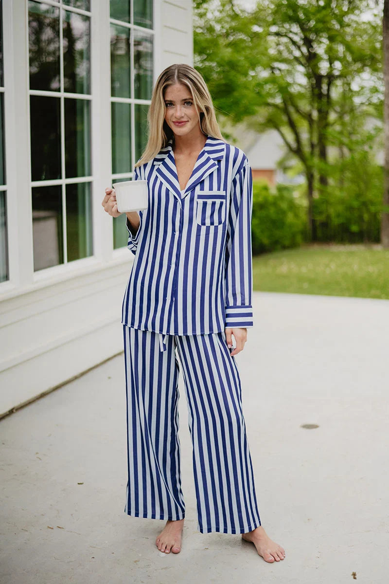 A woman stands barefoot on a patio, smiling while holding a white mug and wearing the Katydid Light Blue and Navy Striped Satin Pajama Matching Set. Lush greenery and a charming house appear in the background.
