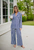 A woman stands barefoot on a patio, smiling while holding a white mug and wearing the Katydid Light Blue and Navy Striped Satin Pajama Matching Set. Lush greenery and a charming house appear in the background.