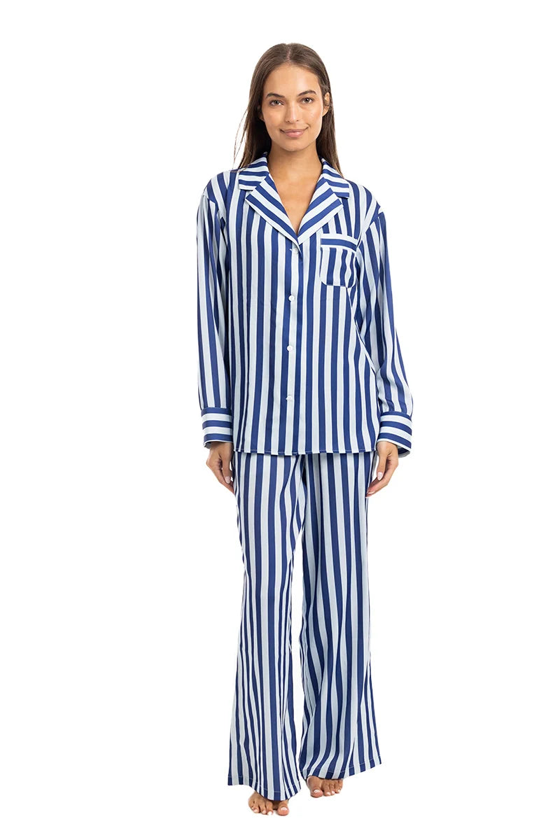 A woman stands facing forward, barefoot against a plain white background, wearing the Katydid Light Blue and Navy Striped Satin Pajama Matching Set featuring a long-sleeve button-up top and matching pants.