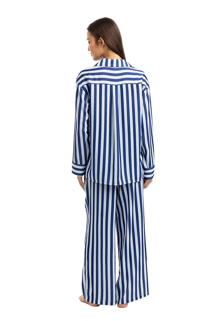 A woman stands barefoot facing away, wearing the Katydid Light Blue and Navy Striped Satin Pajama Matching Set, featuring bold vertical stripes on a long-sleeve top and wide-leg pants against a plain white background.
