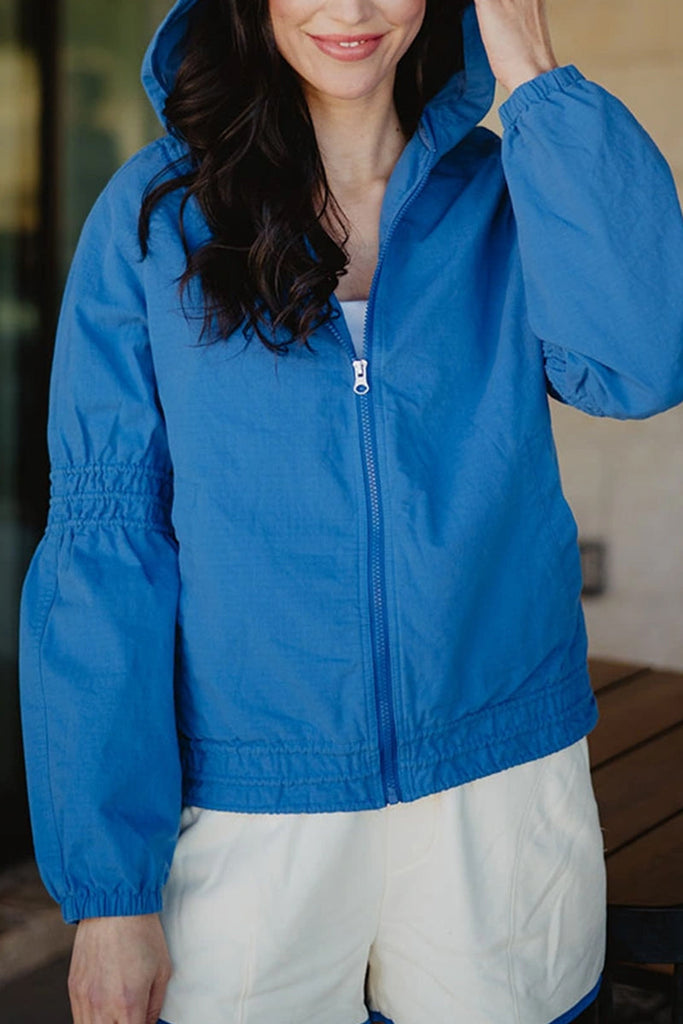 A woman with long dark hair wears the Katydid Blue Lightweight Hooded Jacket and white shorts, standing outdoors with her hand on the hood. The women’s zip-up jacket is casual, and the background is slightly blurred.