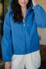 A woman with long dark hair wears the Katydid Blue Lightweight Hooded Jacket and white shorts, standing outdoors with her hand on the hood. The women’s zip-up jacket is casual, and the background is slightly blurred.