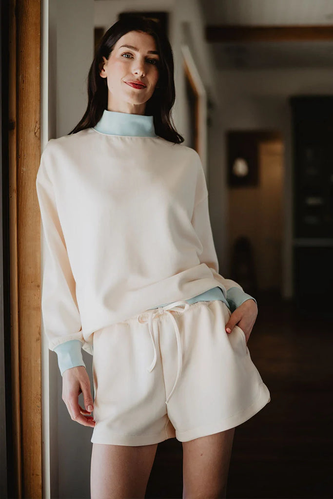 A woman stands indoors smiling, wearing the Katydid Beige  Light Blue Duo Mockneck Matching Set—cream top with light blue cuffs and matching drawstring shorts—with one hand in her pocket.