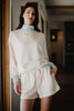 A woman stands indoors smiling, wearing the Katydid Beige  Light Blue Duo Mockneck Matching Set—cream top with light blue cuffs and matching drawstring shorts—with one hand in her pocket.
