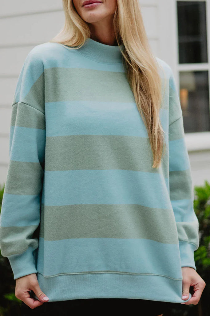 A woman with long blonde hair wears the Katydid Aqua and Green Striped Mockneck Sweatshirt for women—featuring wide, light blue and sage green stripes—while standing outside in front of a white building.
