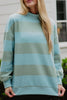A woman with long blonde hair wears the Katydid Aqua and Green Striped Mockneck Sweatshirt for women—featuring wide, light blue and sage green stripes—while standing outside in front of a white building.
