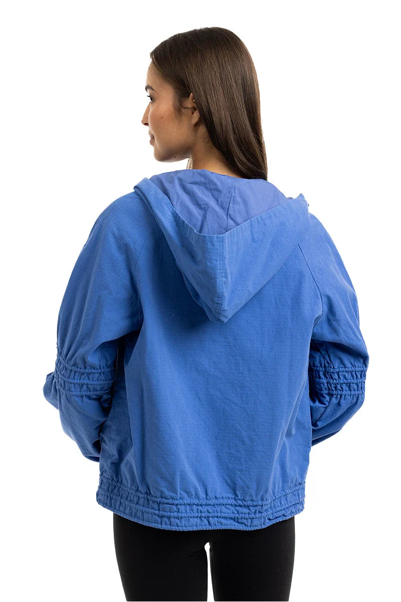 A woman with straight brown hair wears the Katydid Blue Lightweight Hooded Jacket and black leggings, standing with her back to the camera against a white background.