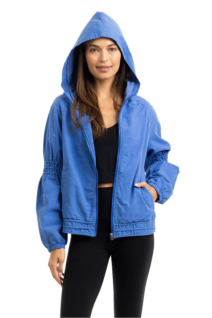 A woman models the Katydid Blue Lightweight Hooded Jacket over a black crop top and leggings, standing with one hand in her pocket and a slight smile against a white background.