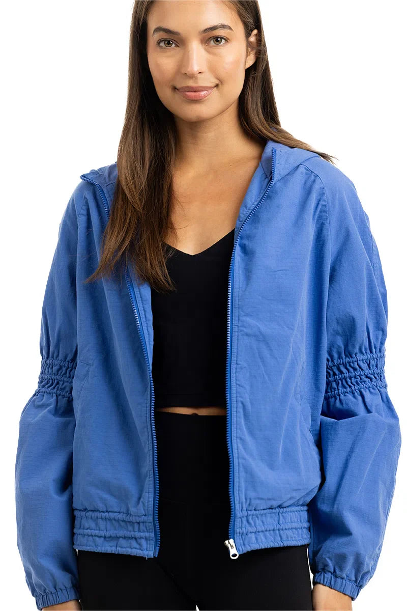 A woman wears the Katydid Blue Lightweight Hooded Jacket over a black crop top, standing facing forward with a slight smile.