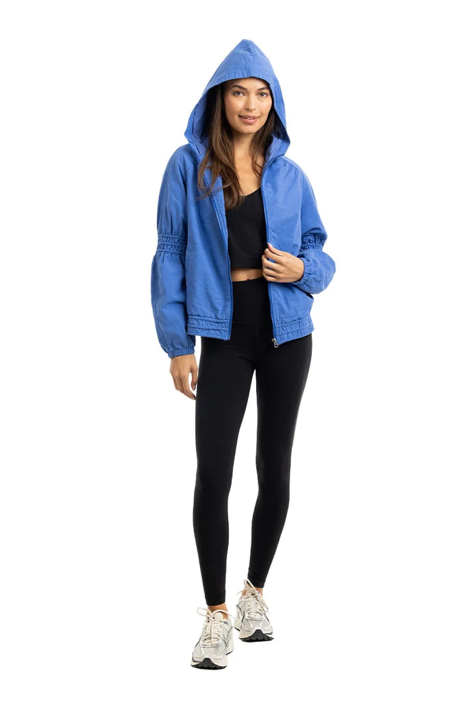 A woman stands facing forward, smiling with one hand in her pocket, wearing a Katydid Blue Lightweight Hooded Jacket over a black crop top and leggings with white sneakers. The jacket adds a stylish touch against the plain white background.