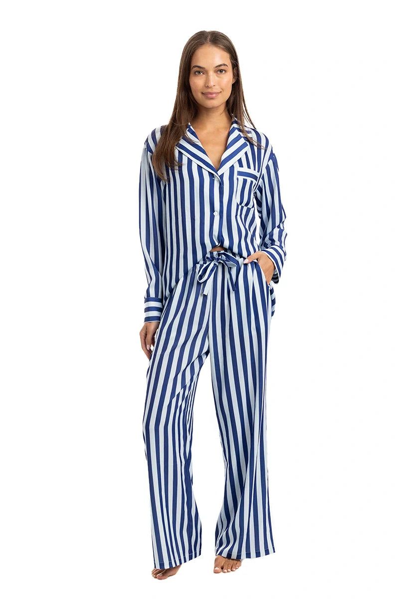 A woman models the Katydid Light Blue and Navy Striped Satin Pajama Matching Set, featuring a button-up long sleeve top and wide-leg pants. She stands barefoot with long straight hair against a white background.