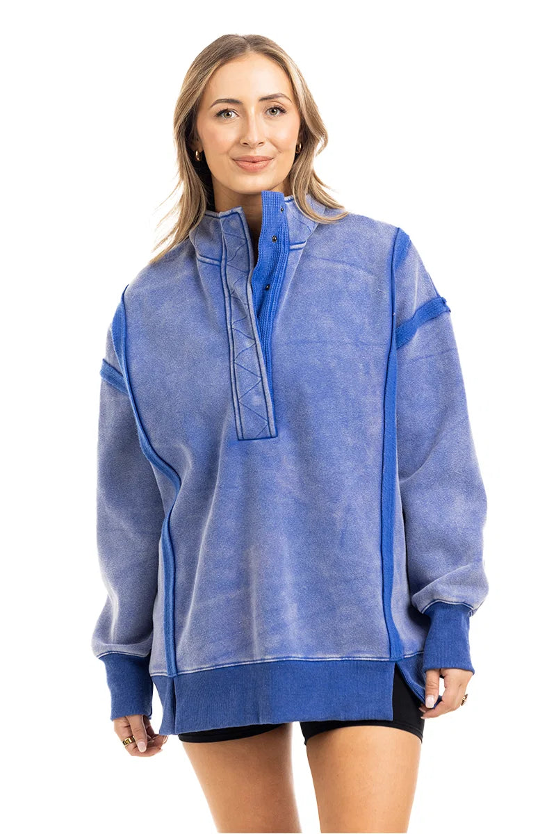 A woman with long hair is smiling against a white background, wearing Katydid’s Blue Vintage Wash Snap-Neck Pullover and black shorts.