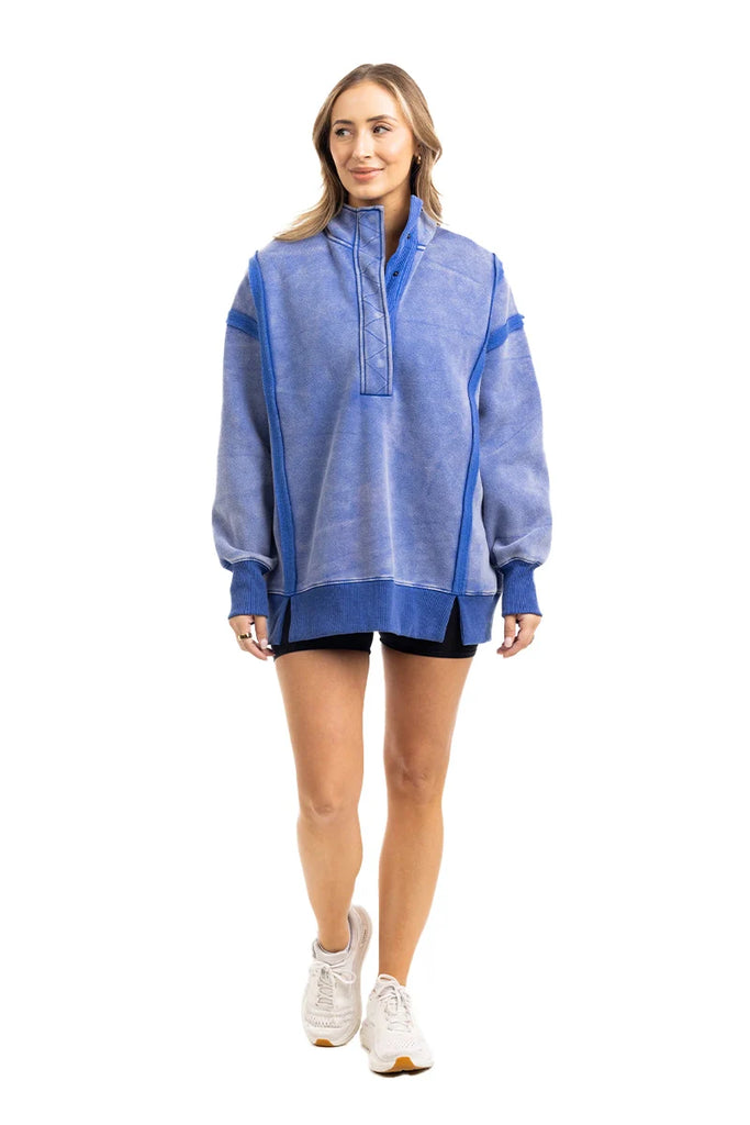 A woman wears the Katydid Blue Vintage Wash Snap-Neck Pullover with black shorts and white sneakers, standing and smiling while looking slightly to the side against a white background.