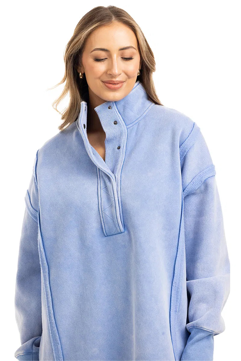 A woman with long, light brown hair wears the Katydid Light Blue Vintage Wash Snap-Neck Pullover. The cozy, loose-fitting top complements her gentle smile as she stands against a white background with her eyes softly closed.