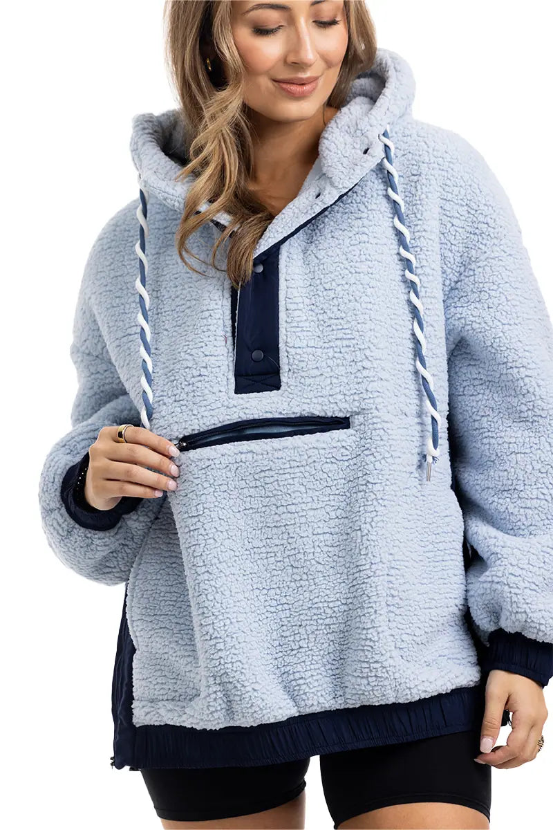 A woman smiles and looks down, wearing the Katydid Light Blue Two-Toned Sherpa Fleece Pullover with navy accents and a front zipper pocket. Her long, wavy hair complements the cozy top, styled effortlessly with black shorts.