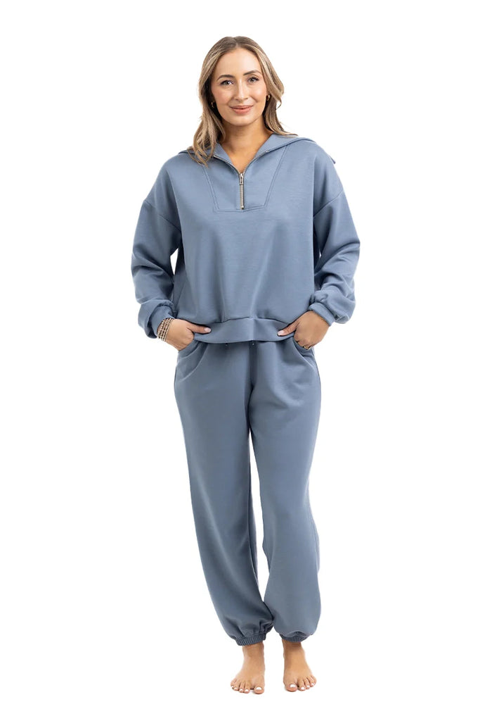 A woman stands barefoot, smiling, with her hands in her pockets, wearing the Katydid Steel Blue Matching Sets Womens Pants—featuring a zippered pullover top and jogger-style pants—on a white background.