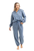 A woman wearing the Katydid Steel Blue Matching Sets Womens Pants—a loose-fitting, long-sleeve zip-up sweatshirt with drawstring pants—smiles barefoot against a white background.