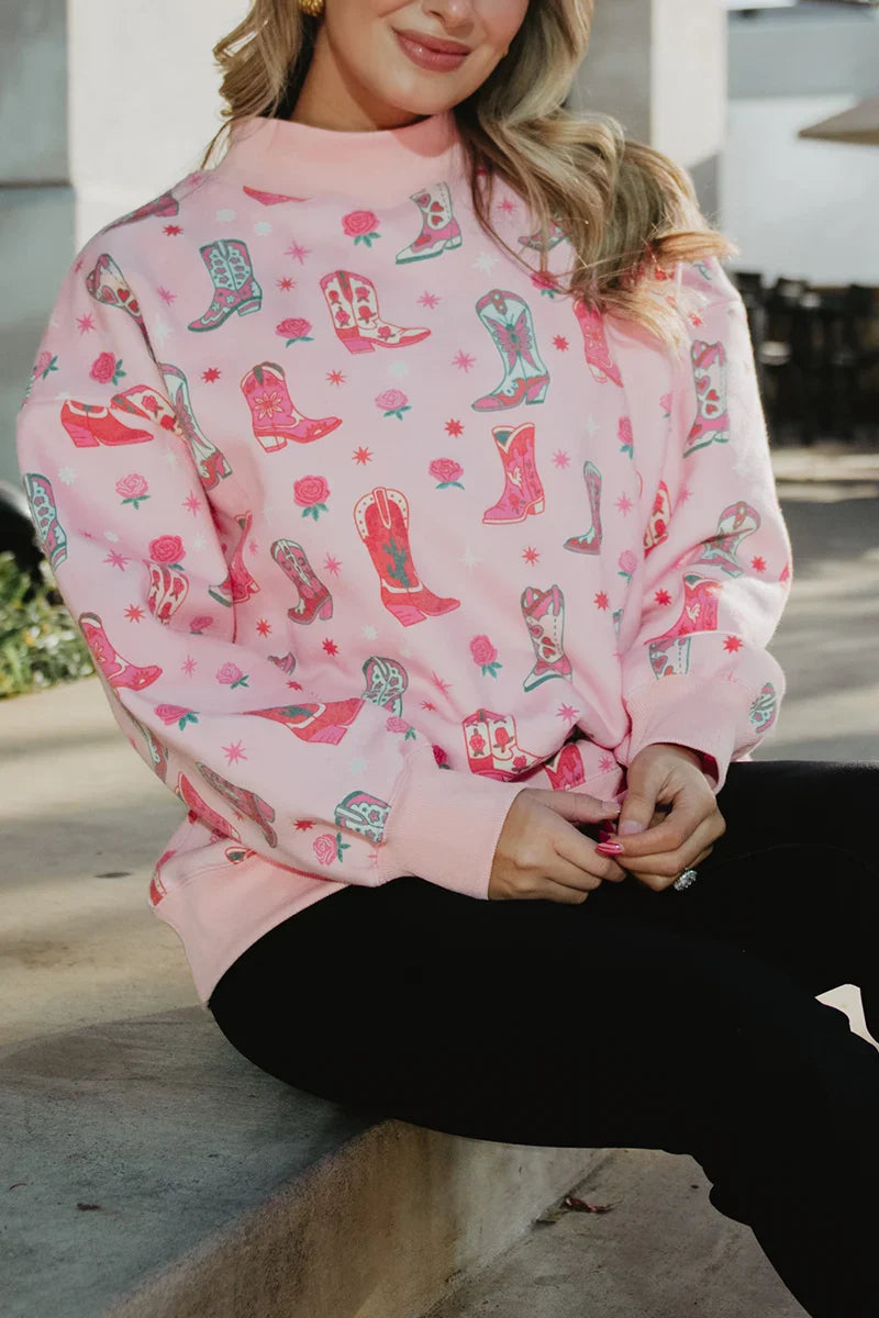 A woman sits on a concrete bench wearing the Katydid Boots  Roses Printed Sweatshirt, which features colorful cowboy boots and floral patterns, paired with black pants.
