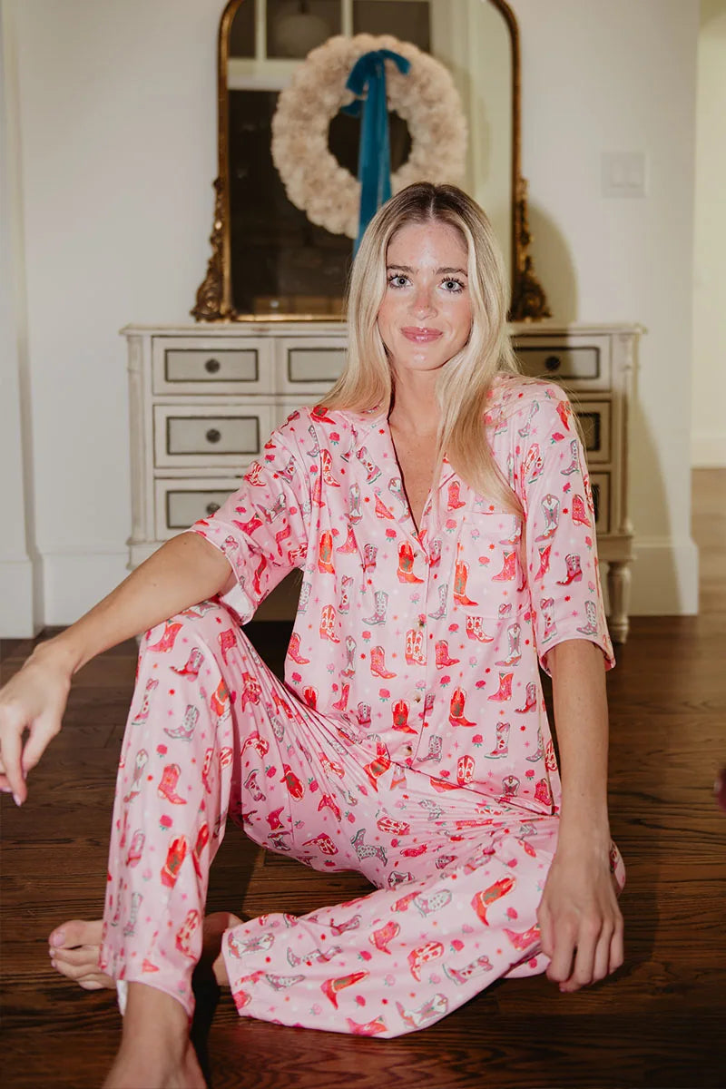 A woman with long blonde hair sits on a wooden floor, wearing the Katydid Womens Boots  Roses PJ Set. She is in front of a white dresser with a mirror, decorated with a wreath and blue ribbon.