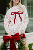 A smiling woman wears the Katydid Bow Patch Reindeer Mockneck Sweatshirt, featuring red bow patches, paired with a red skirt. She stands outdoors holding festive greenery with a large red bow, her blonde hair flowing loosely.