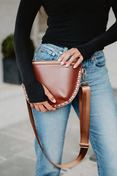 A person in a black long-sleeve top and blue jeans wears the Katydid Brown Gold-Studded Crossbody Sling Bag with gold studs and a wide strap at their waist—perfect for everyday style.