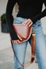 A person in a black long-sleeve top and blue jeans wears the Katydid Brown Gold-Studded Crossbody Sling Bag with gold studs and a wide strap at their waist—perfect for everyday style.