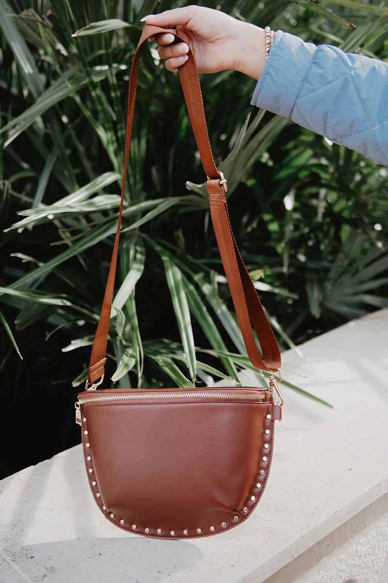 A hand holds up the Katydid Brown Gold-Studded Crossbody Sling Bag, featuring gold studs along the edges, set against green leafy plants.