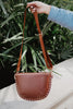 A hand holds up the Katydid Brown Gold-Studded Crossbody Sling Bag, featuring gold studs along the edges, set against green leafy plants.