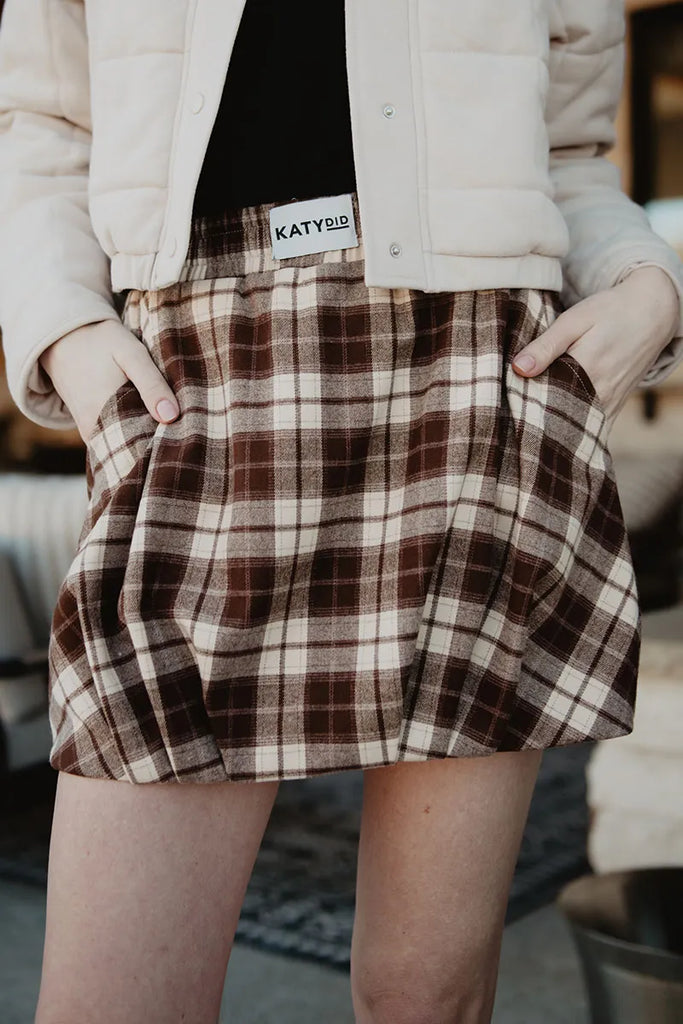 A person wears a beige jacket and a Brown Plaid Bubble Hem Skirt with hands in pockets. The skirt’s waistband features a Katydid label.