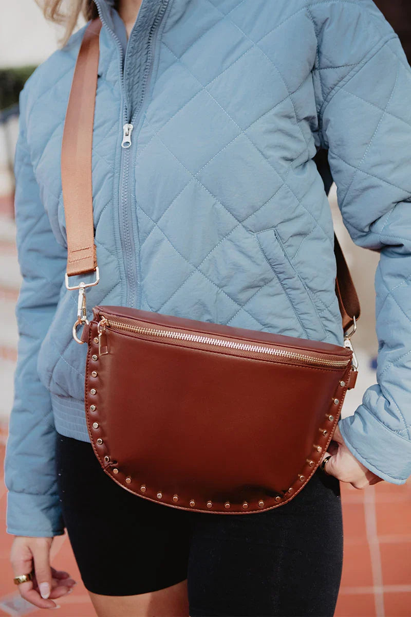 A person in a light blue quilted jacket and black shorts carries the Katydid Brown Gold-Studded Crossbody Sling Bag across their chest, featuring gold studs and a wide tan strap—an ideal accessory to elevate everyday looks.