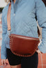 A person in a light blue quilted jacket and black shorts carries the Katydid Brown Gold-Studded Crossbody Sling Bag across their chest, featuring gold studs and a wide tan strap—an ideal accessory to elevate everyday looks.