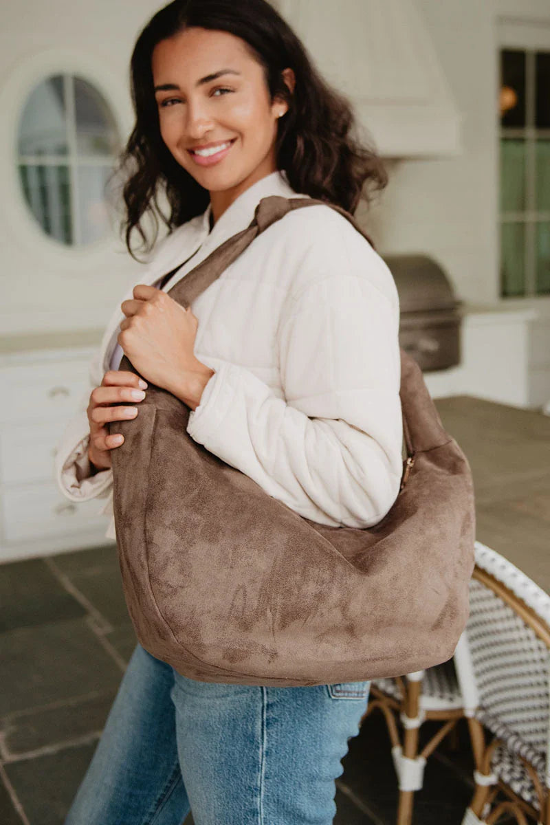 A woman with wavy dark hair, in a light jacket and blue jeans, smiles while holding the Katydid Coffee Brown Suede Totes in a bright, modern indoor space.
