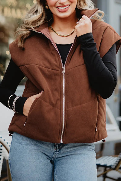 A woman smiles outdoors, wearing the Katydid Brown Corduroy Quilted Vest over a black long-sleeve shirt, blue jeans, and gold jewelry—one hand in her pocket and the other on her vest—for perfect fall style.